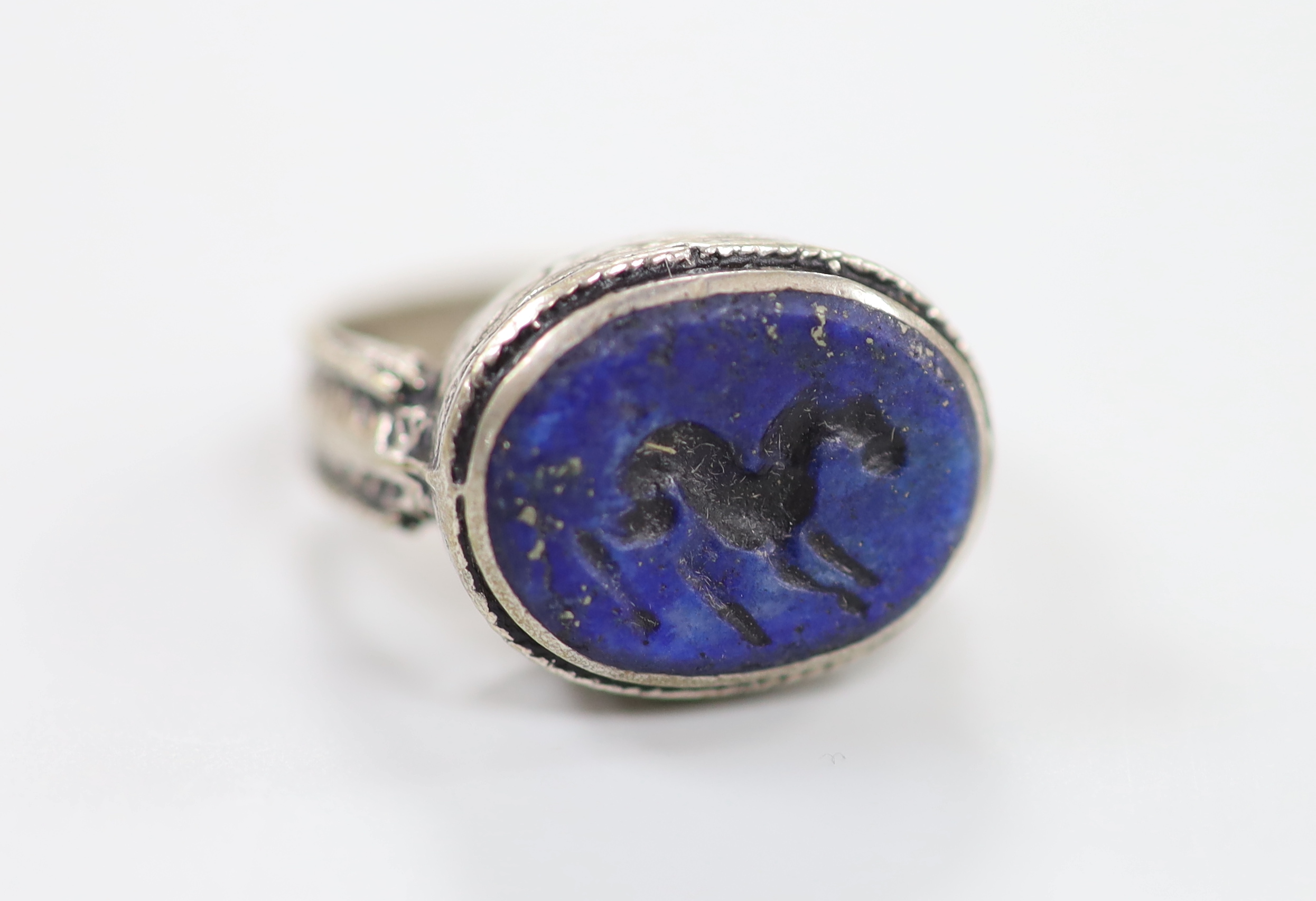 An engraved white metal and intaglio lapis lazuli set oval ring, the matrix carved with a horse?, size R/S, gross weight 7.4 grams.